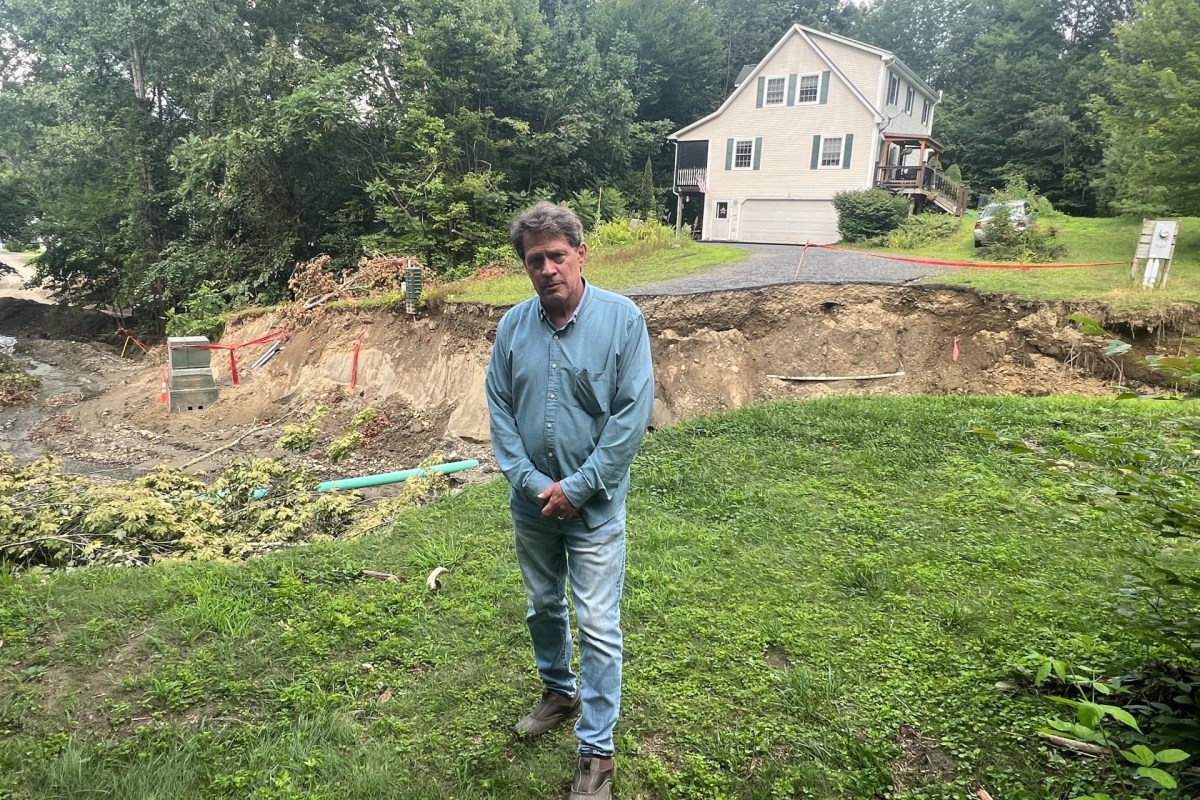 A man stands on grass with his hands clasped, in front of a house near a large eroded area, partly barricaded with safety fencing.