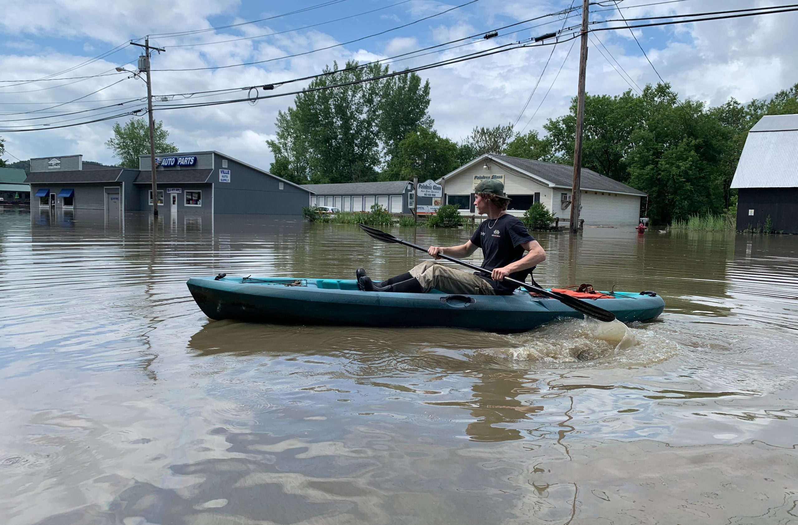A person paddles a kayak down a flooded street lined with partially submerged buildings under a cloudy sky.