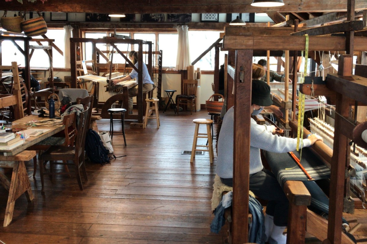 A workshop with several wooden weaving looms where people are working on textiles. The room has wooden floors and beams, and is furnished with various chairs and tables.