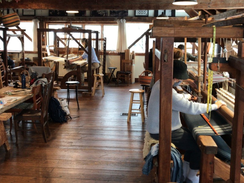 A workshop with several wooden weaving looms where people are working on textiles. The room has wooden floors and beams, and is furnished with various chairs and tables.