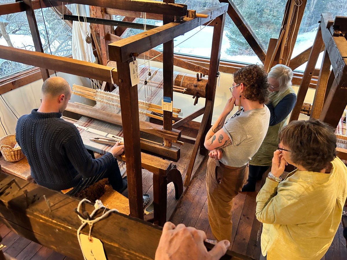 A person weaves on a large wooden loom while four others observe in a workshop setting with large windows.