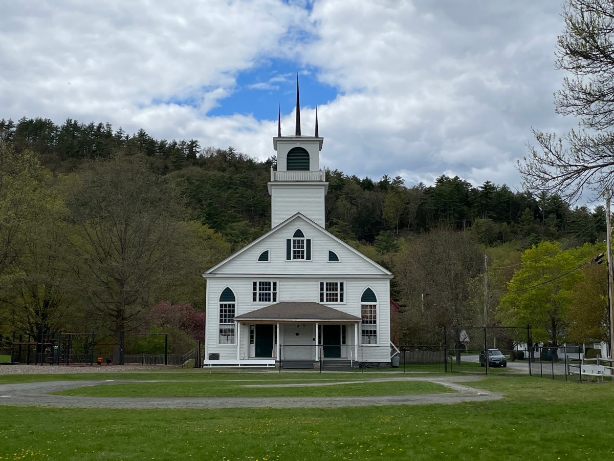 A white, wooden church with a tall steeple stands in front of a forested hill under a partly cloudy sky. There is a grassy field with a circular path in the foreground.
