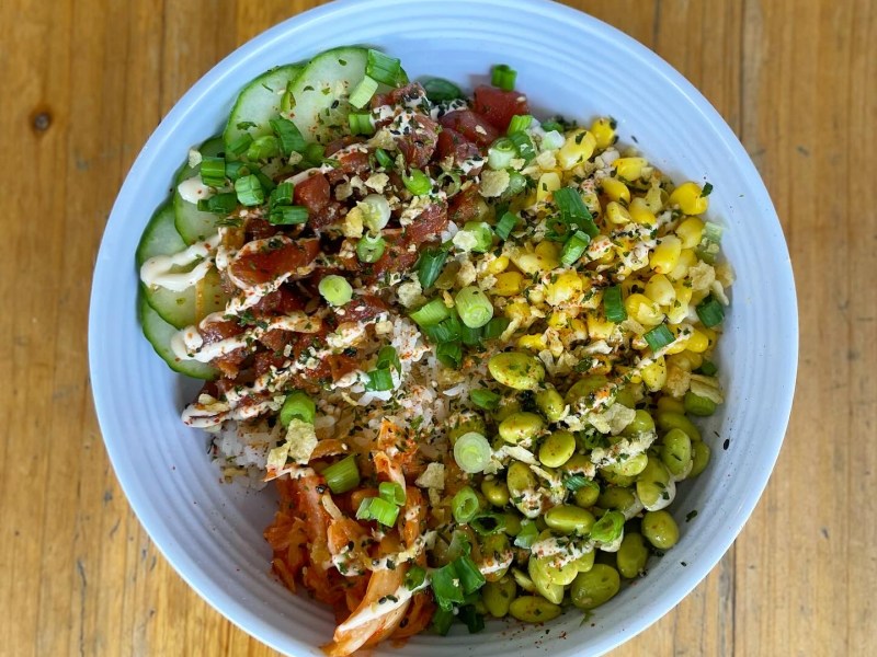 A bowl of food with rice, topped with sliced cucumber, corn, edamame, shredded carrots, green onions, sesame seeds, and a drizzle of white sauce, all on a wooden table.