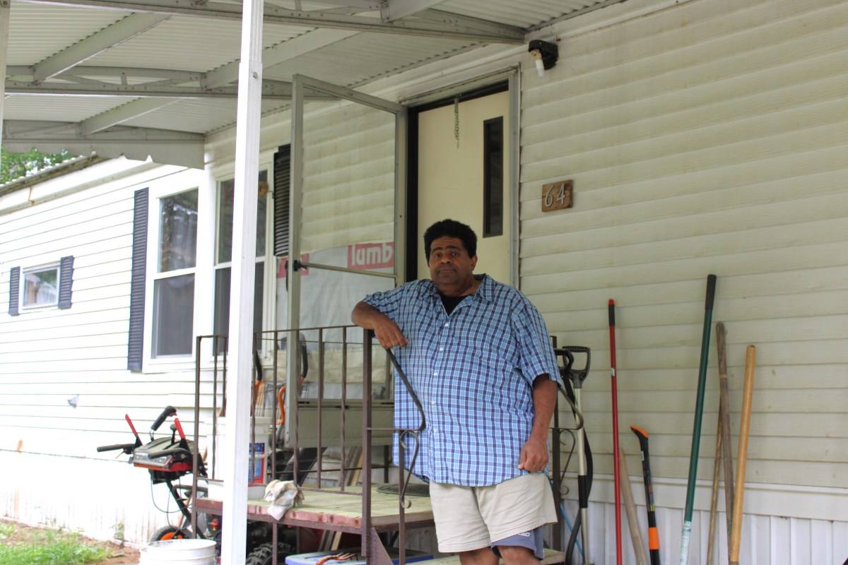A man in a plaid shirt and shorts stands on the porch of a mobile home with a screen door, surrounded by garden tools and equipment.