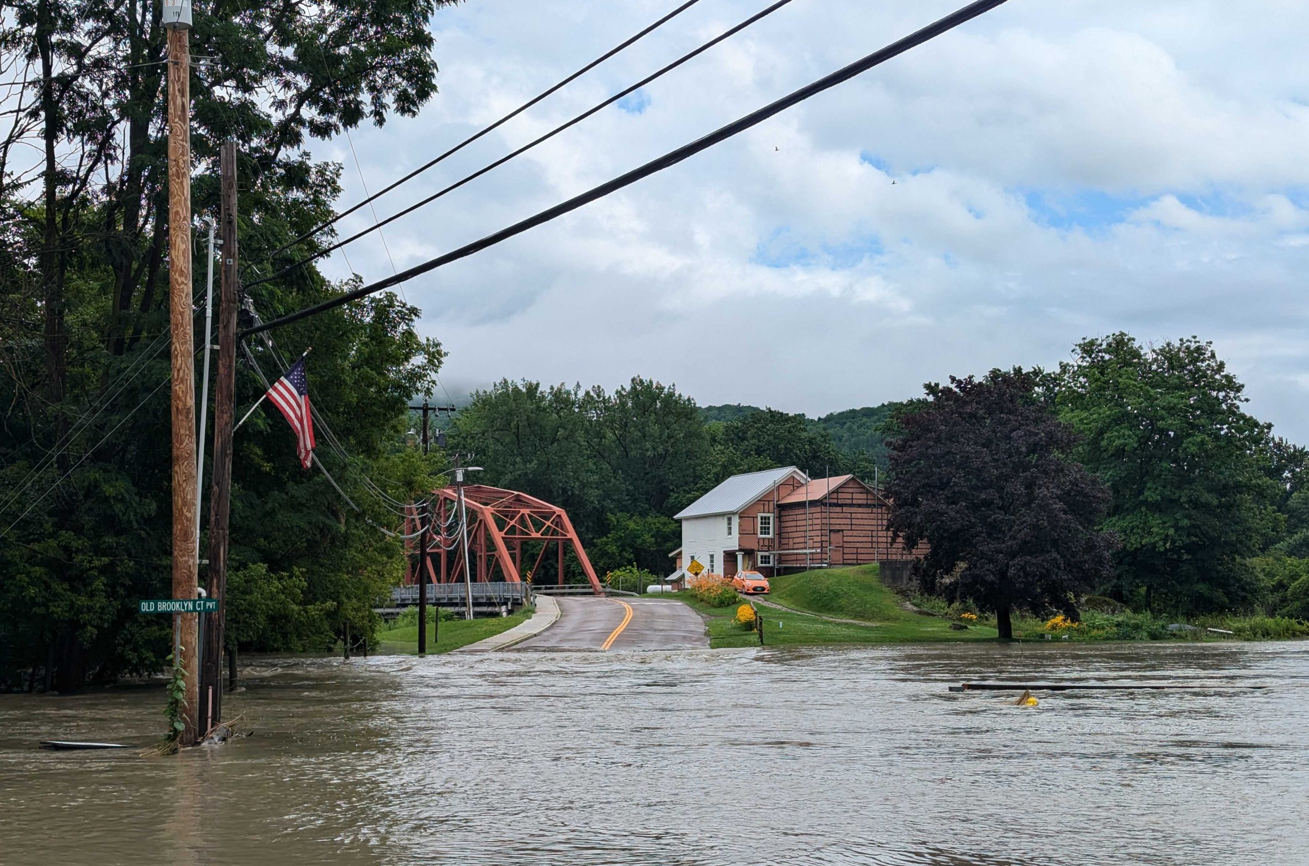 A flooded street blocks access to a red truss bridge and a house. A partially submerged road sign and an American flag on a utility pole are visible in the foreground.