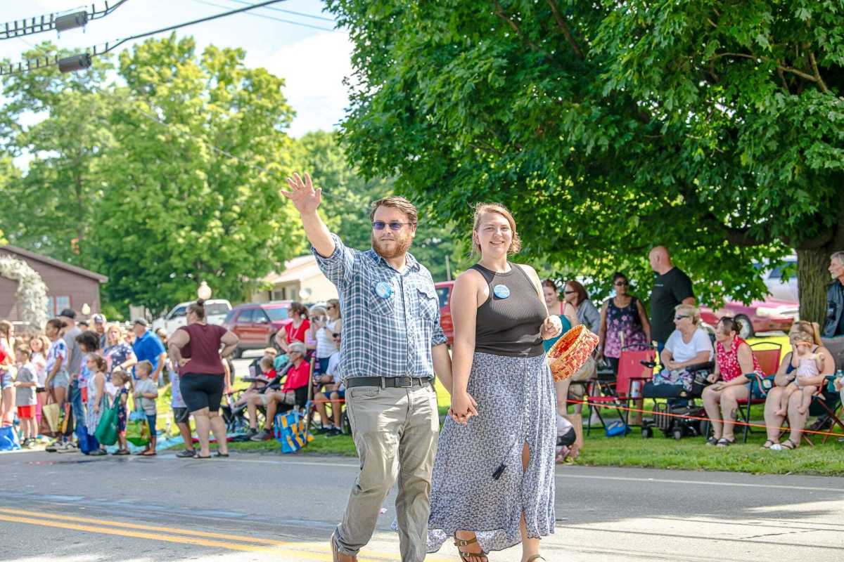 A man and a woman walk down a street, waving and smiling at a parade. They are surrounded by people sitting and standing along the roadside.