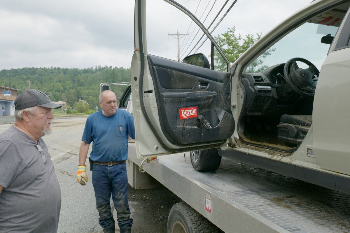 Two men stand beside an open car door on a tow truck platform. The car door has a "Bernie" sticker near the bottom. Trees and buildings are visible in the background.