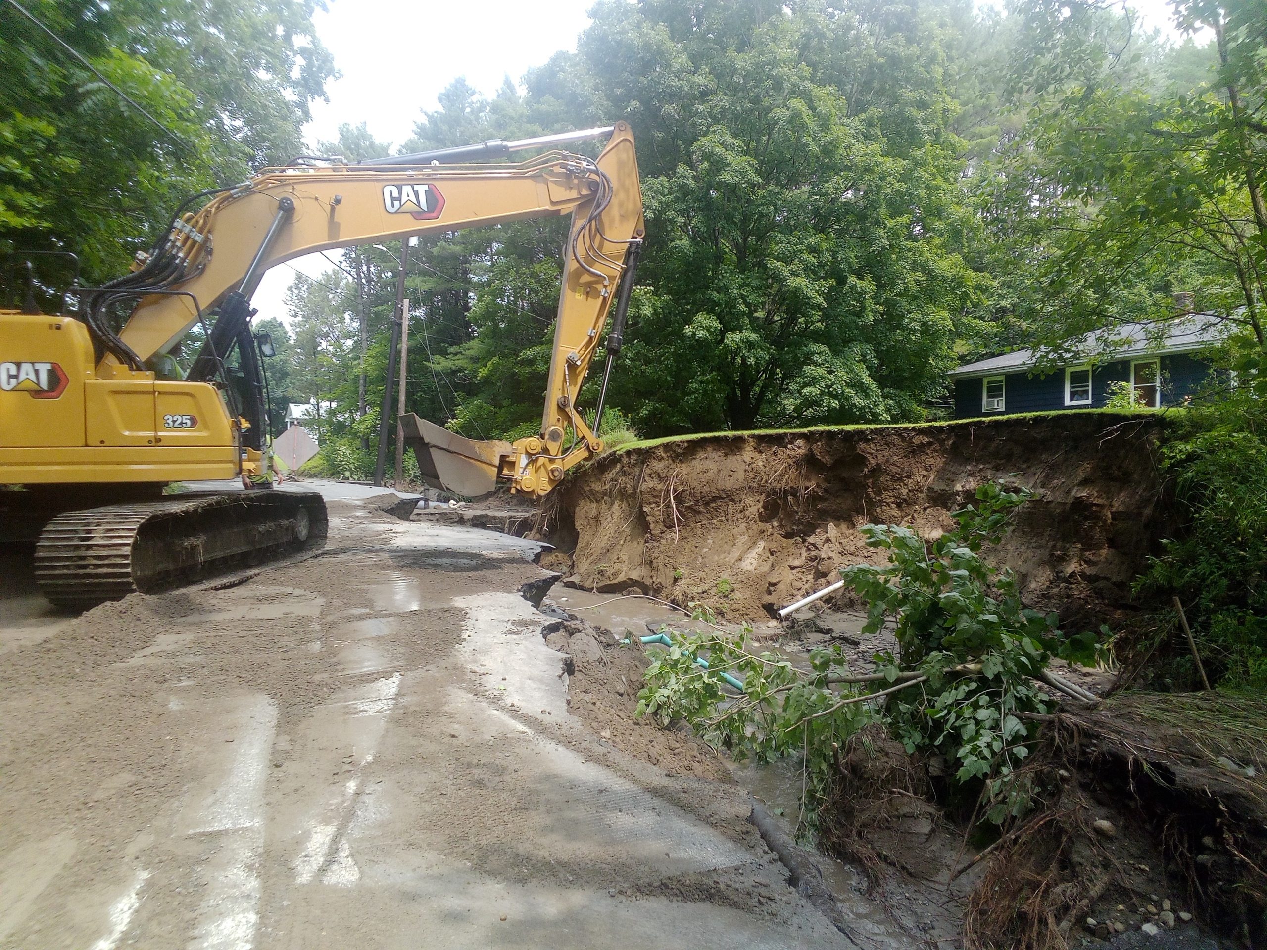 A yellow excavator works on a road next to a large landslide that has caused significant erosion near a house surrounded by trees.