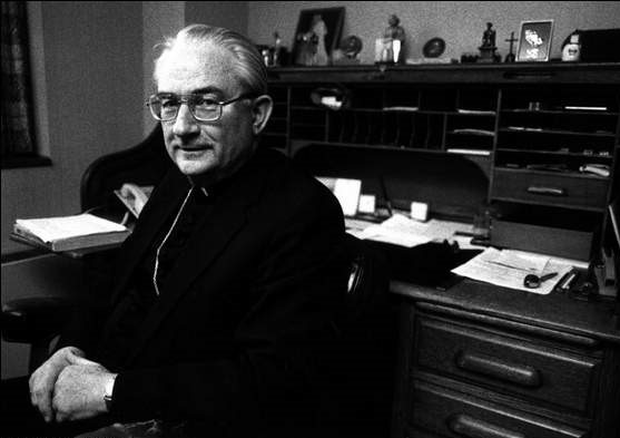 An elderly man with glasses and wearing a clerical collar sits beside a cluttered wooden desk in a dimly lit office.