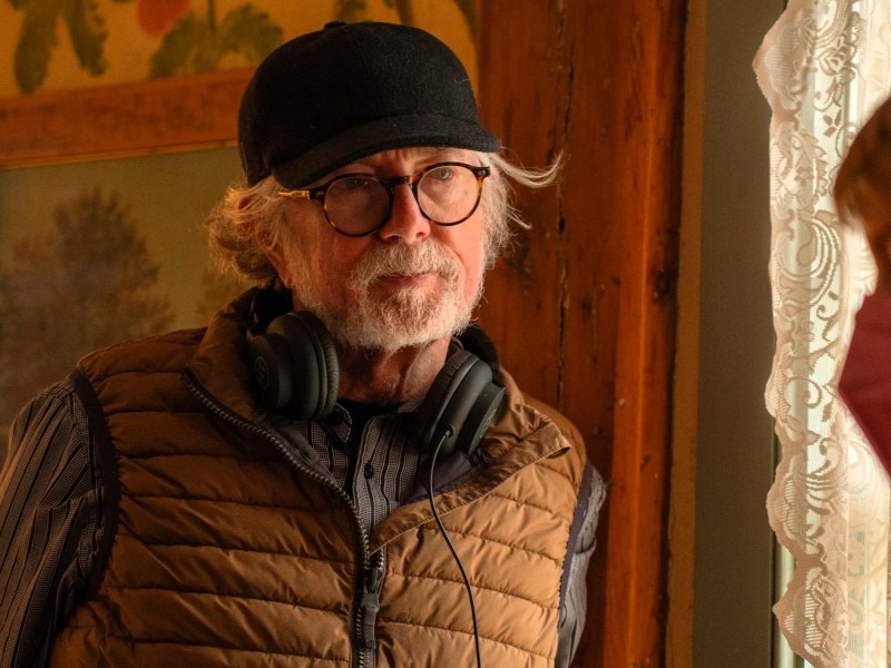 An older man with glasses and a black hat stands by a window with lace curtains, wearing headphones around his neck and a brown puffer vest.