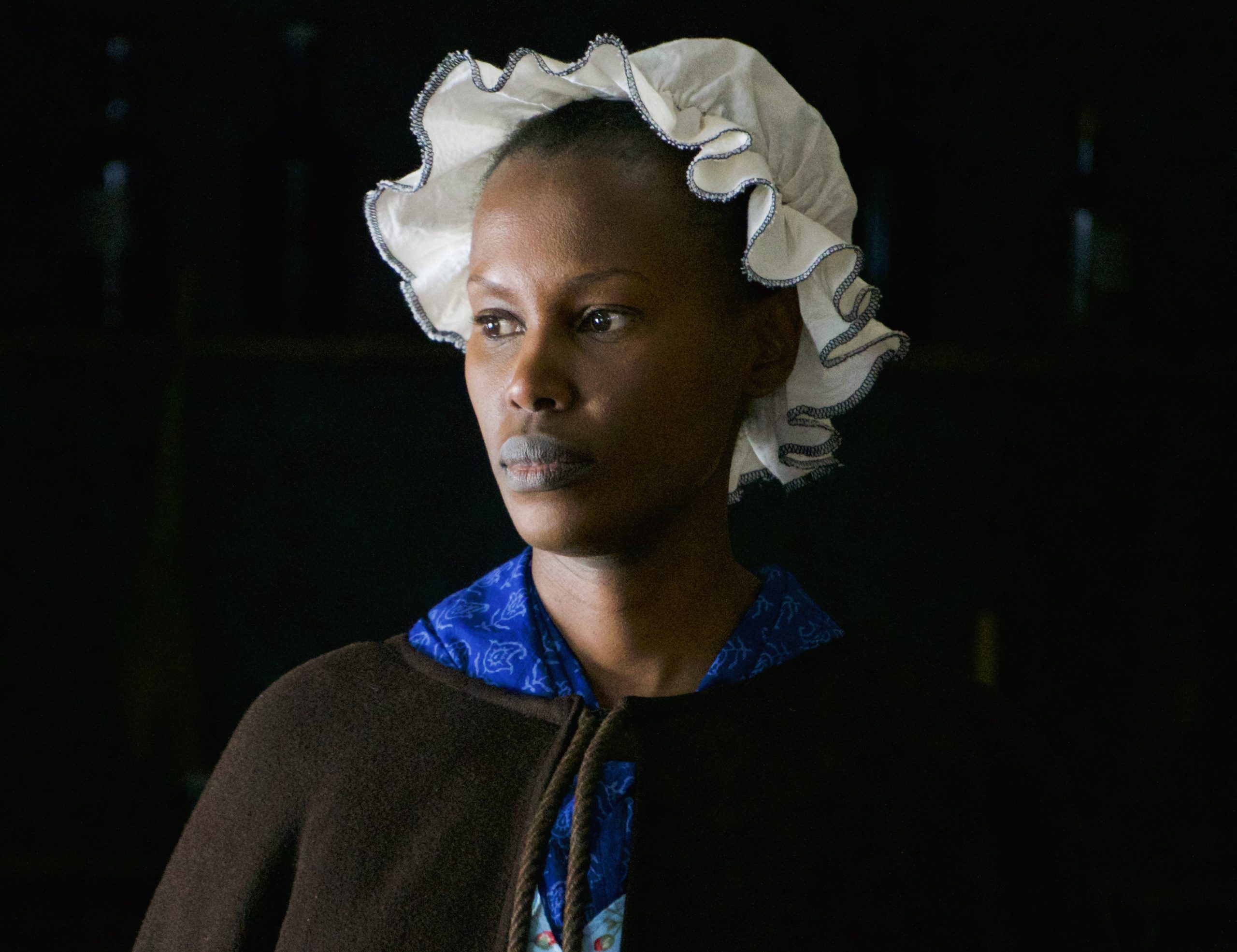 A person wearing a white bonnet and a dark shawl looks off to the side against a dark background.