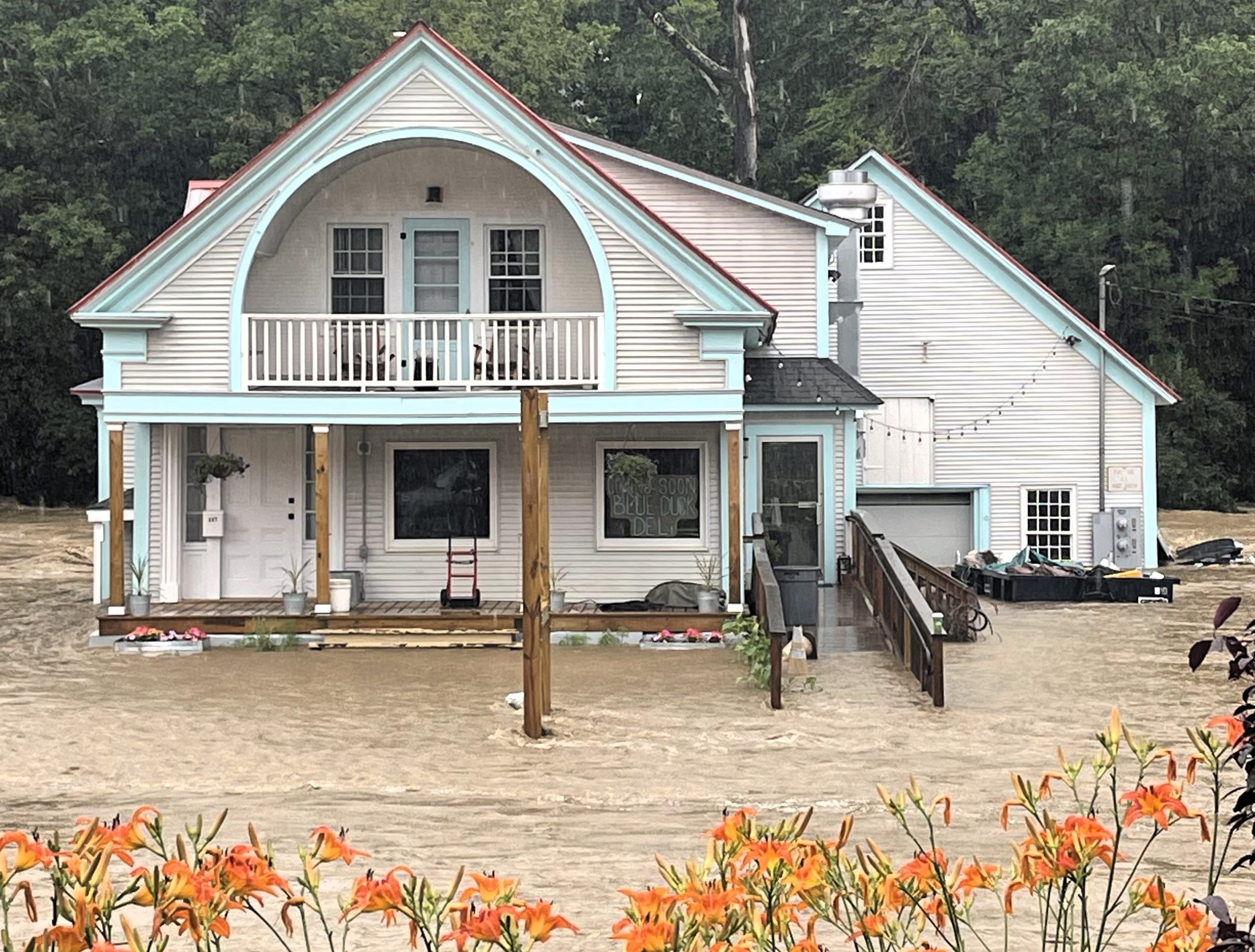 A house surrounded by floodwater with sandbags at the entrance. The yard is submerged, and the water level reaches the base of the house. Orange flowers are in the foreground.