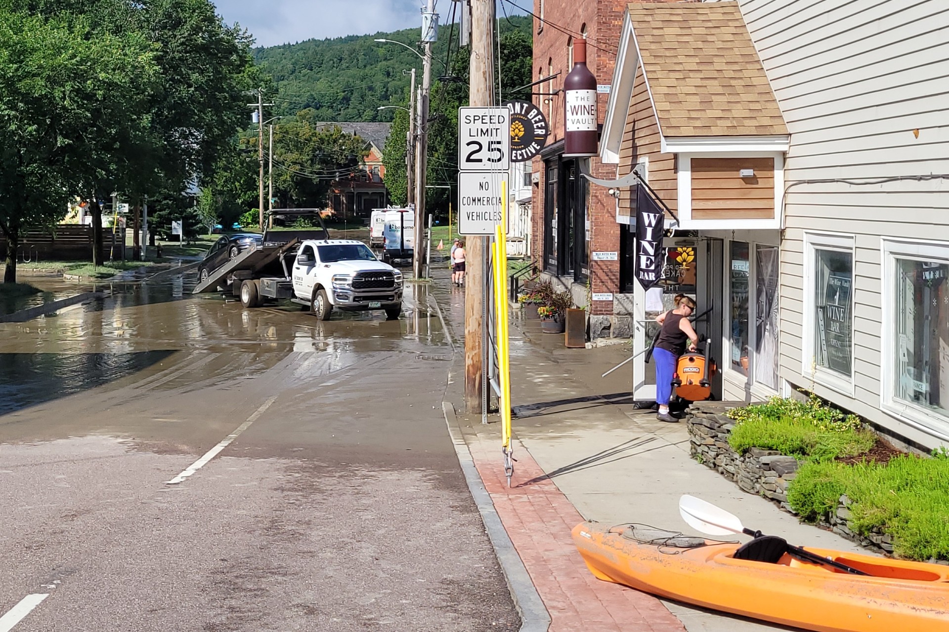 A street is flooded with water, and a tow truck is assisting a white truck. A person stands in front of a building labeled "Wine," and a kayak is on the sidewalk.