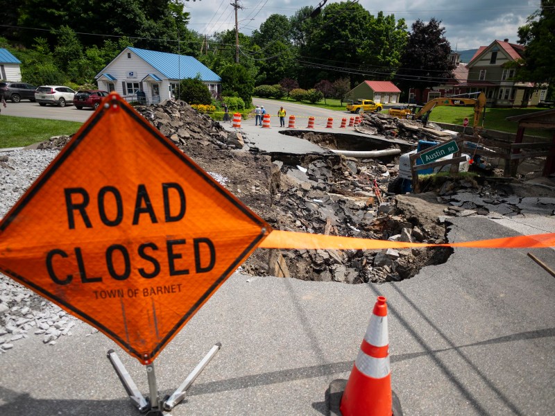 Road closed sign and orange cones block access to a street severely damaged by a sinkhole in a residential area.