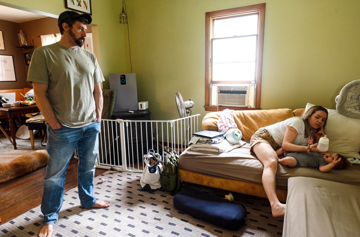 A man stands in a room while a woman feeds a baby with a bottle on a sofa. The room has a baby gate, window AC unit, and various items scattered around.