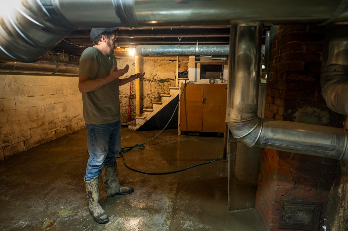 A person in muddy boots gestures while standing in a flooded basement with exposed pipes and a staircase in the background.