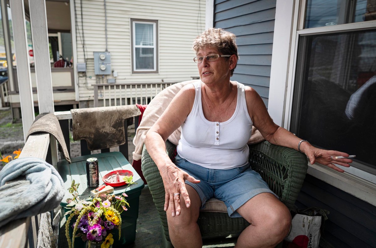 A woman in a white tank top and denim shorts sits on a porch. There is a small table with a drink, plate, and flowers next to her. She looks to the side with her arm resting on the chair.