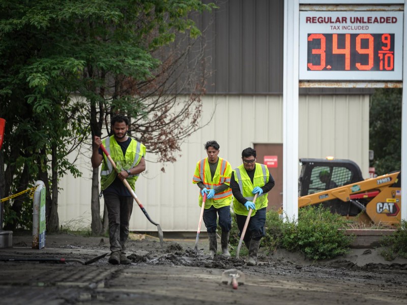 Three workers in yellow safety vests clean up mud on a street near a gas station with a sign showing regular unleaded gasoline priced at $3.49 per gallon.