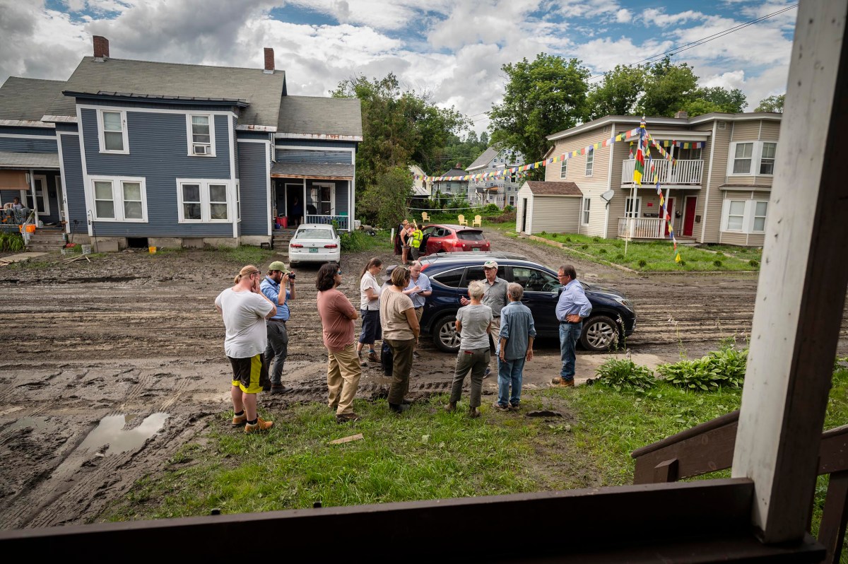A group of people stands in a muddy street between two houses, with several cars parked nearby. Multicolored flags are strung across the street, and clouds cover the sky.