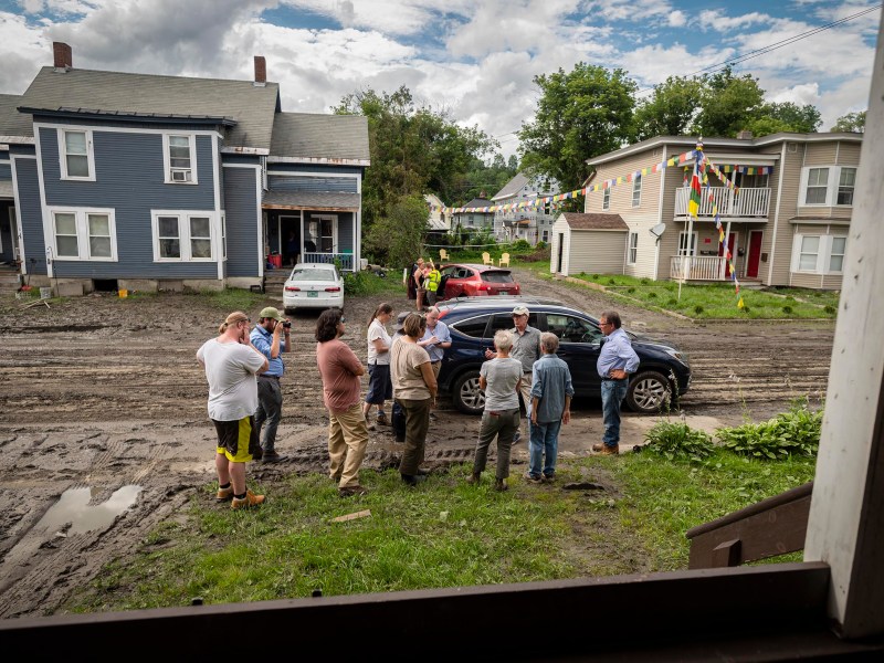A group of people stands in a muddy street between two houses, with several cars parked nearby. Multicolored flags are strung across the street, and clouds cover the sky.