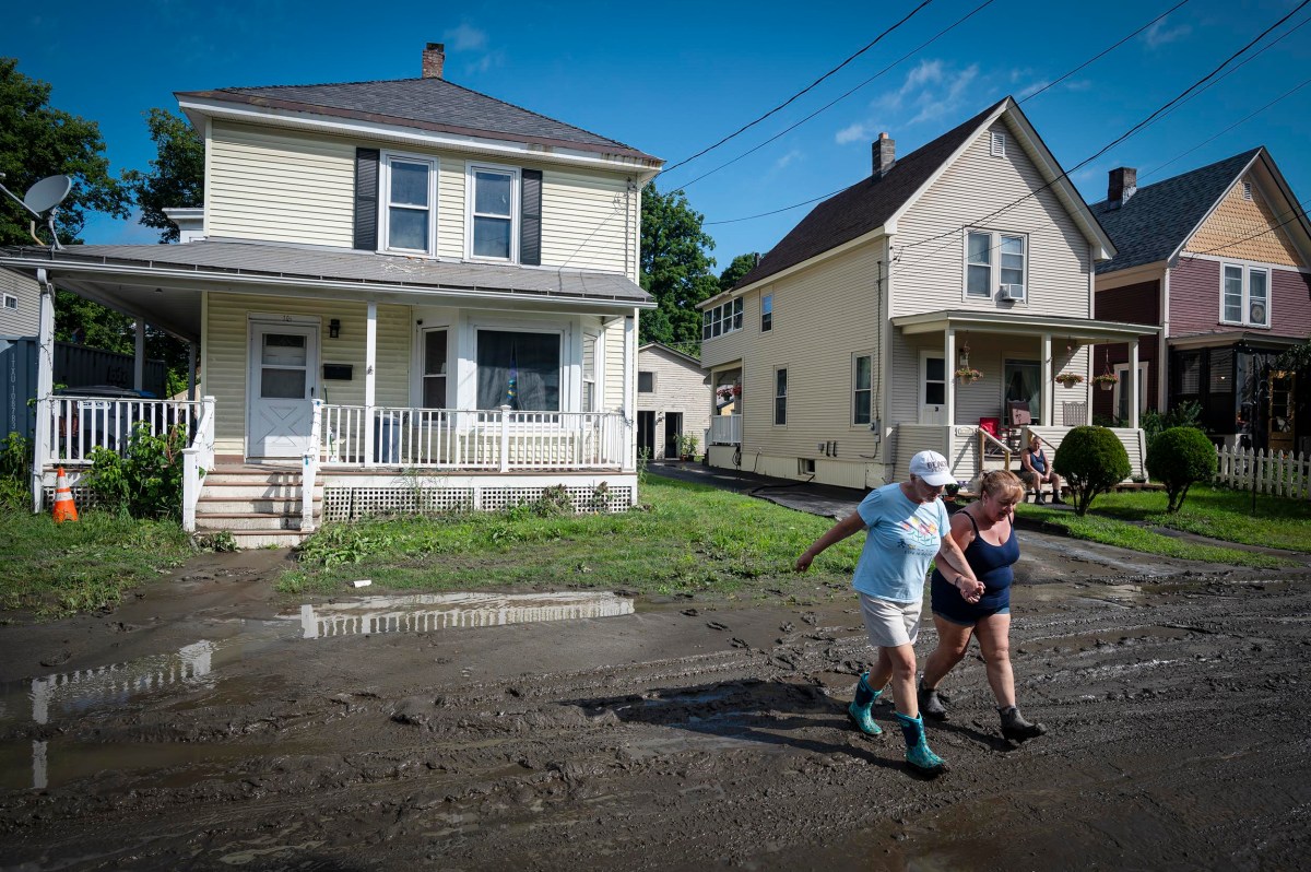 Two people walk through a muddy street in a residential neighborhood with houses in the background, after what appears to be a heavy rain or flooding.