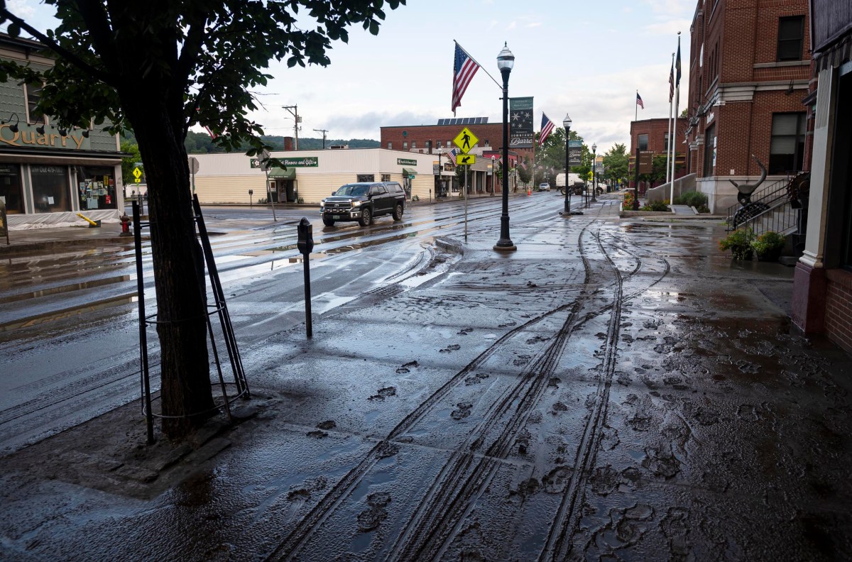 A muddy street with tire and foot tracks following a rainstorm. An SUV drives in the distance, and American flags hang from poles along the sidewalk lined with buildings.