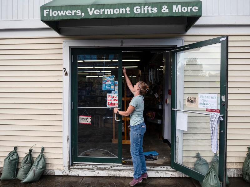 A person adjusts a sign on the glass door of a shop named "Flowers, Vermont Gifts & More." Sandbags are positioned outside the entrance, and an open side door displays a "Please Use Other Entrance" sign.