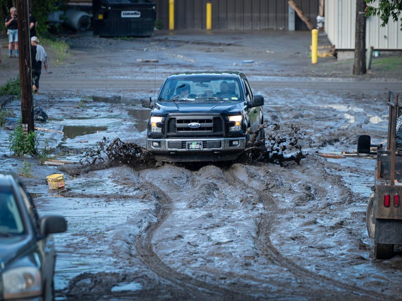 A black pickup truck drives through a large muddy puddle on a dirt road, splashing water and mud. People and other vehicles are visible in the background.