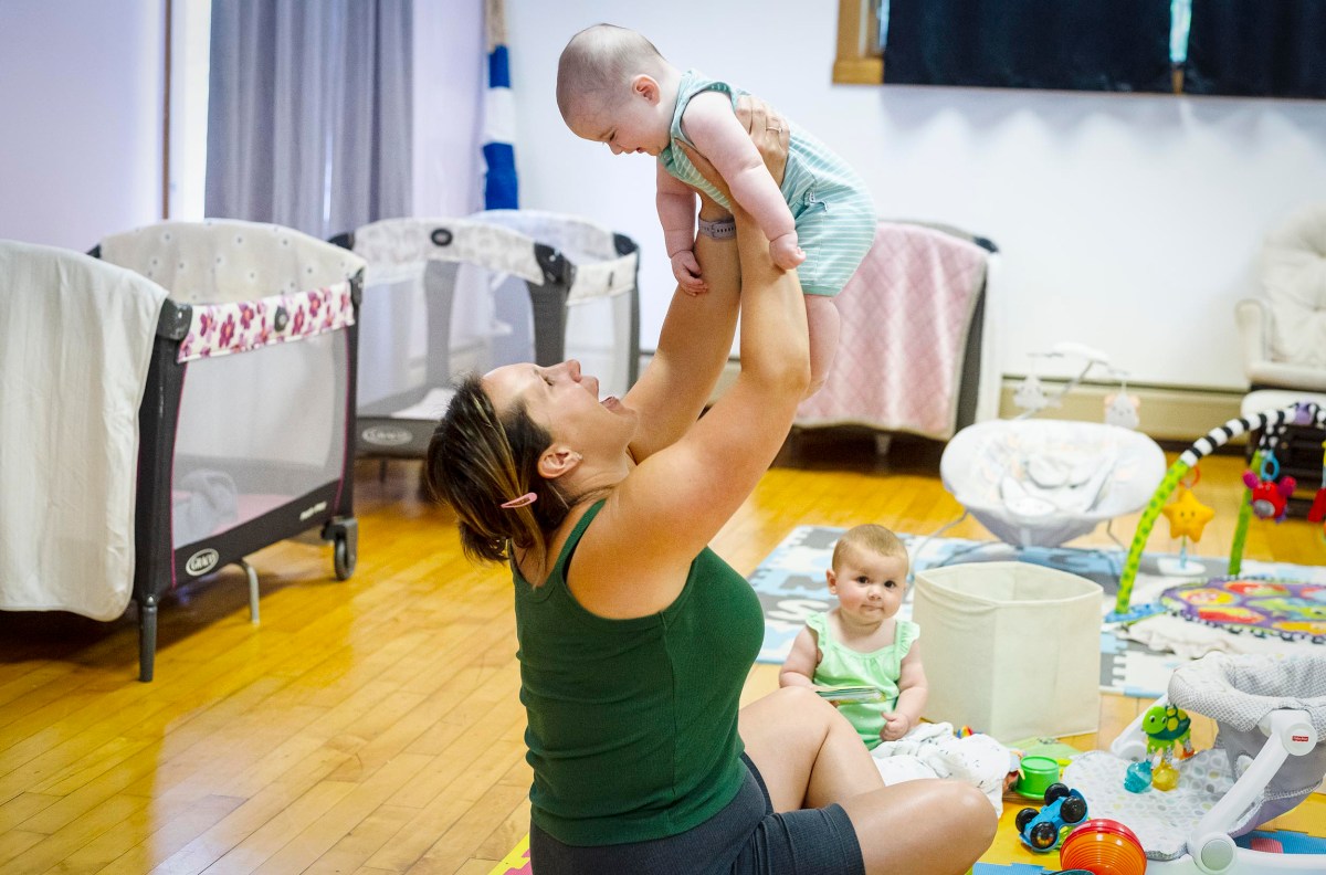 An adult lifts a baby in the air while another baby sits nearby among toys on the floor of a room with cribs and playpens.