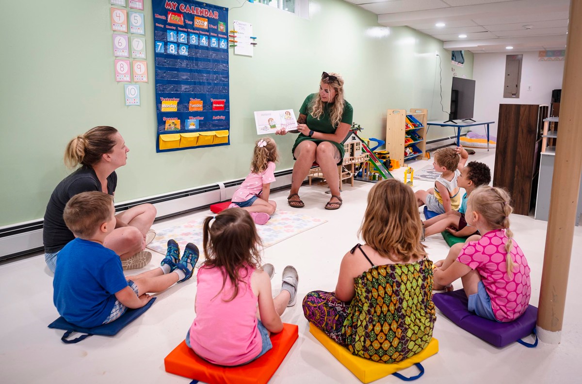 A teacher is reading a book to a group of young children seated on colorful mats in a classroom. The wall behind them features a calendar, alphabet chart, and various educational materials.
