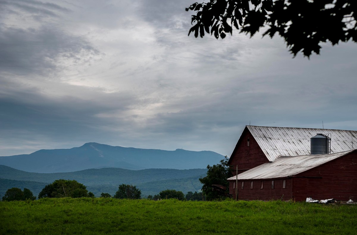 A red barn sits in a green field under a cloudy sky, with a mountain range in the background.