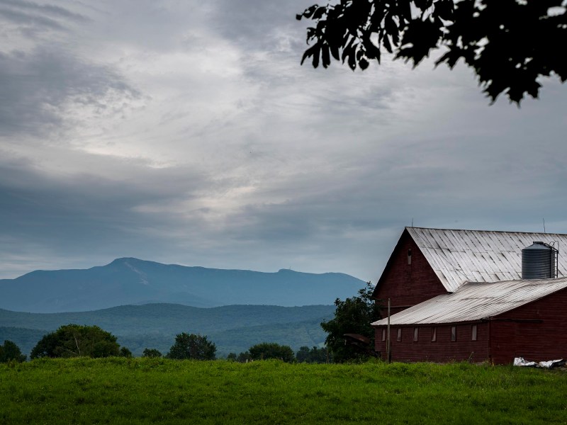 A red barn sits in a green field under a cloudy sky, with a mountain range in the background.