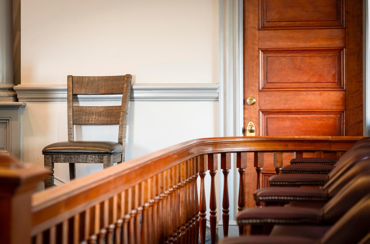 A wooden chair sits next to a closed wooden door in a courtroom, with empty jury seats and a wooden railing in the foreground.