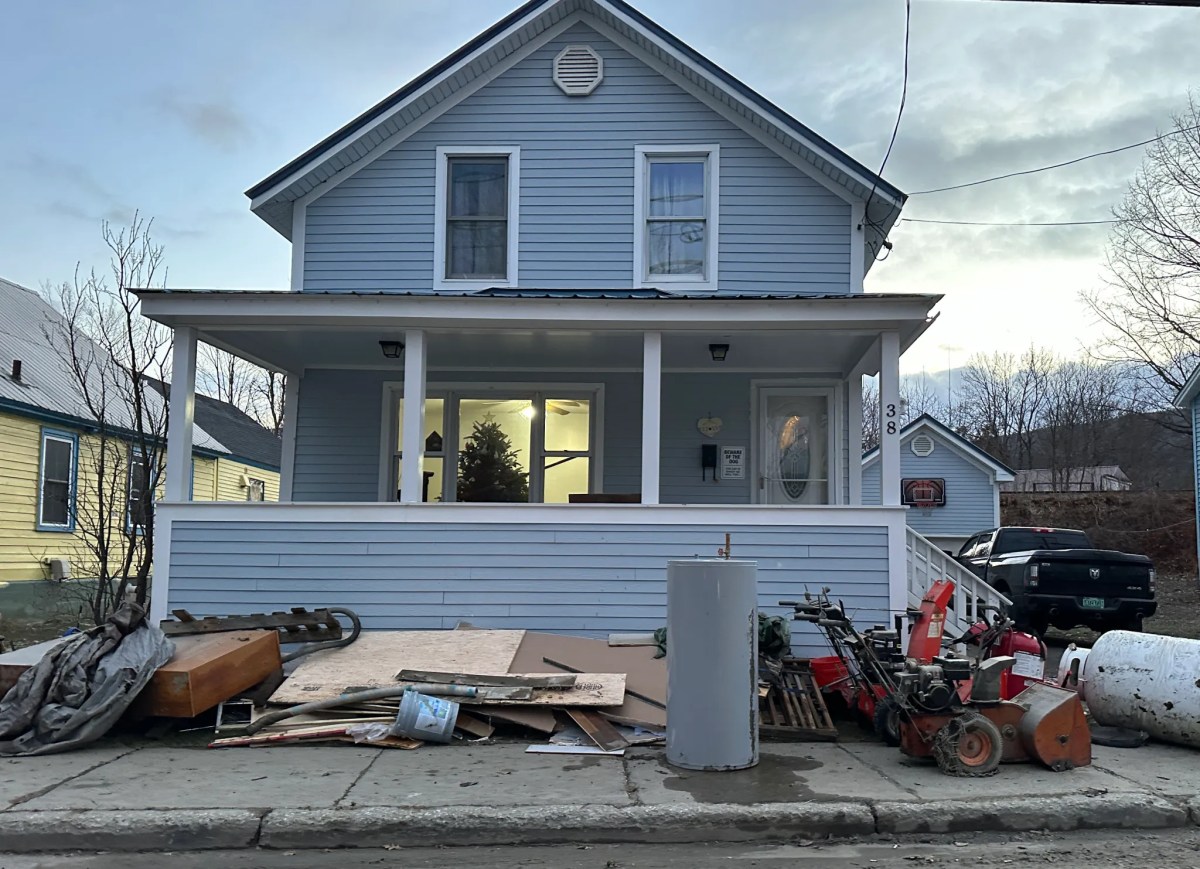A blue house with a front porch has various items and debris, including furniture, boards, and tools, scattered along the sidewalk in front of it.
