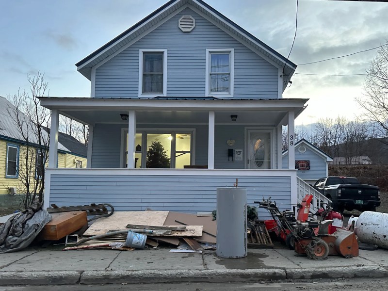A blue house with a front porch has various items and debris, including furniture, boards, and tools, scattered along the sidewalk in front of it.