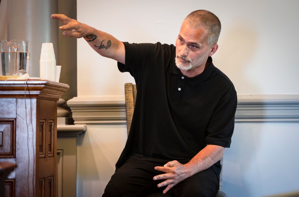 A man with a gray beard and a tattoo on his arm speaks while pointing in a direction, seated near a wooden podium with plastic cups and a water pitcher.
