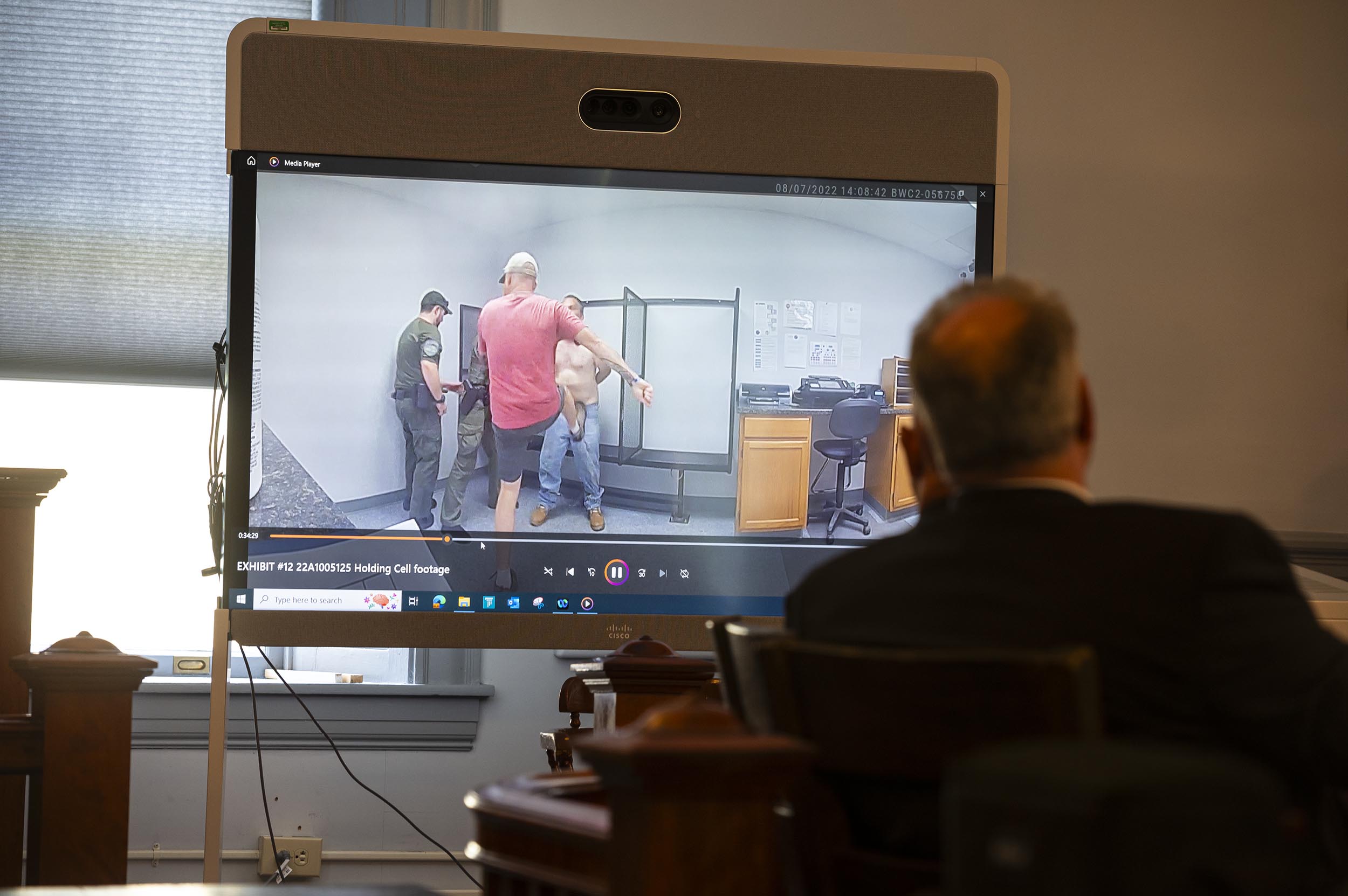A monitor in a courtroom displays security footage of a person being detained by two officers. A person sits in the foreground, watching the screen attentively.