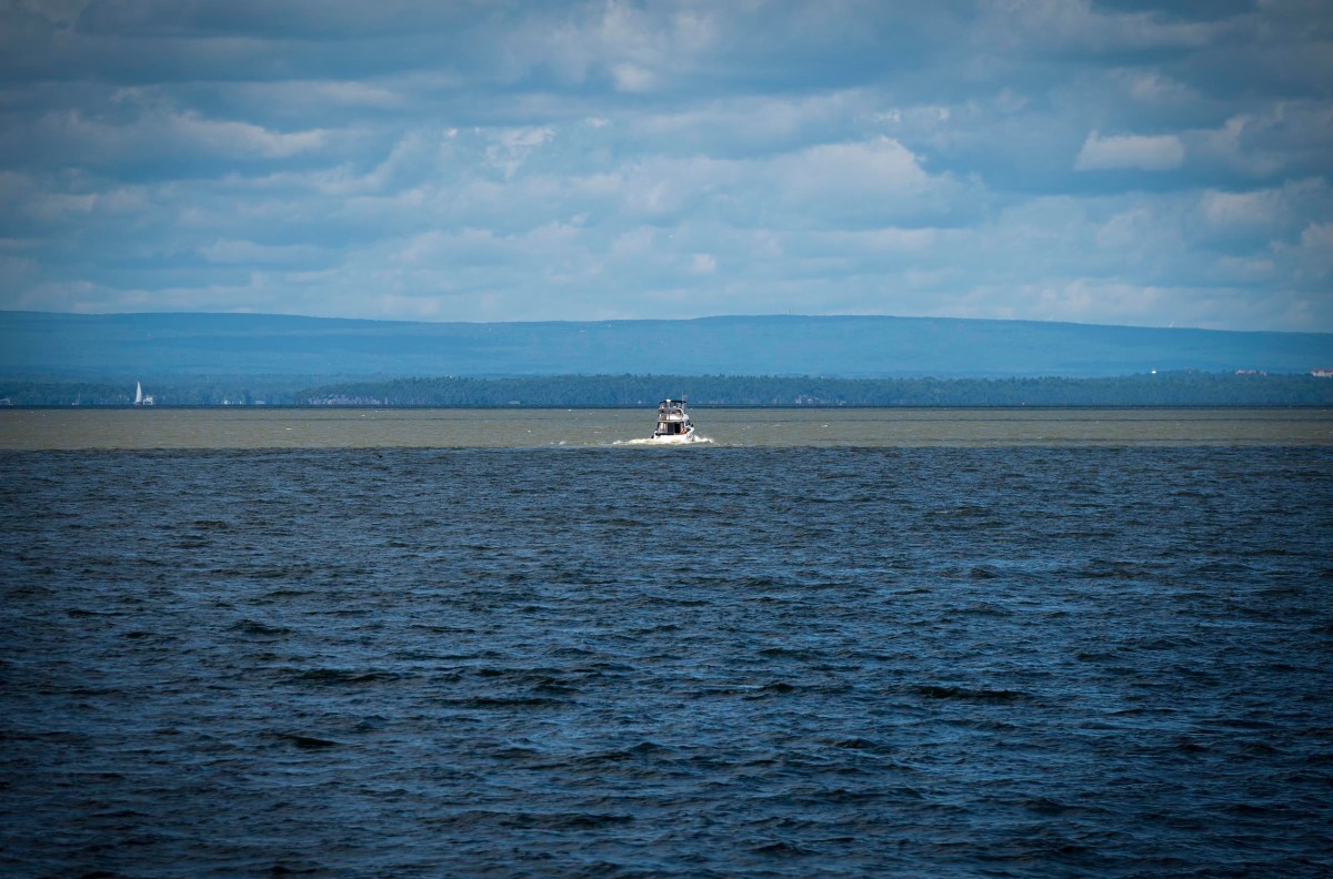 A boat in the distance travels across a large body of water, under a cloudy sky with land visible on the horizon.