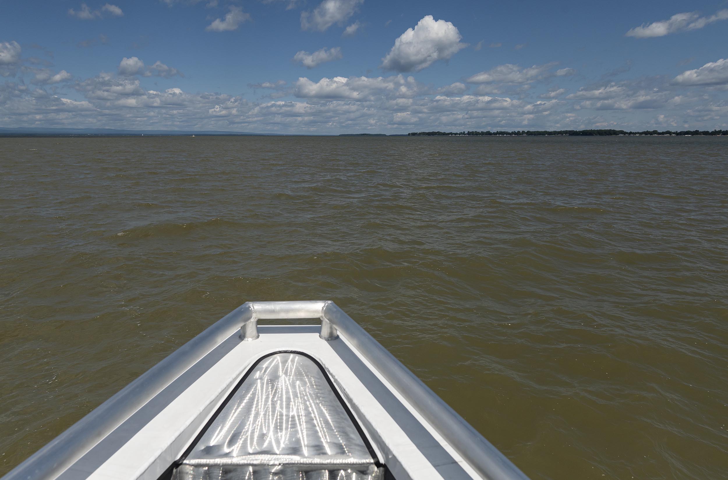 The bow of a boat is shown heading across a body of brownish water under a partly cloudy sky with distant land visible on the horizon.