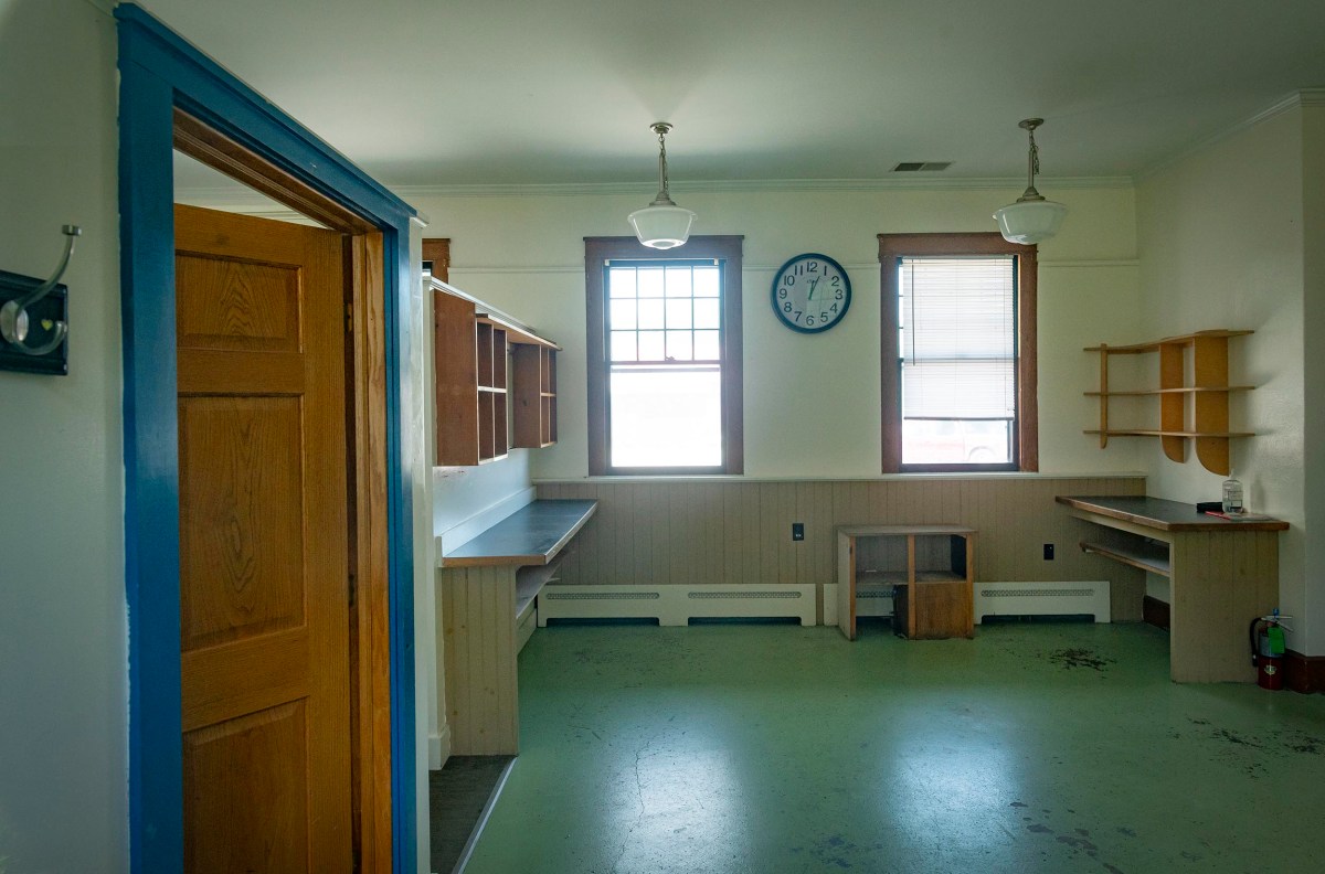 A sparsely furnished room with two windows, a clock on the wall, shelves, a desk, and wooden door. The floor is green with some marks, and there are light fixtures hanging from the ceiling.