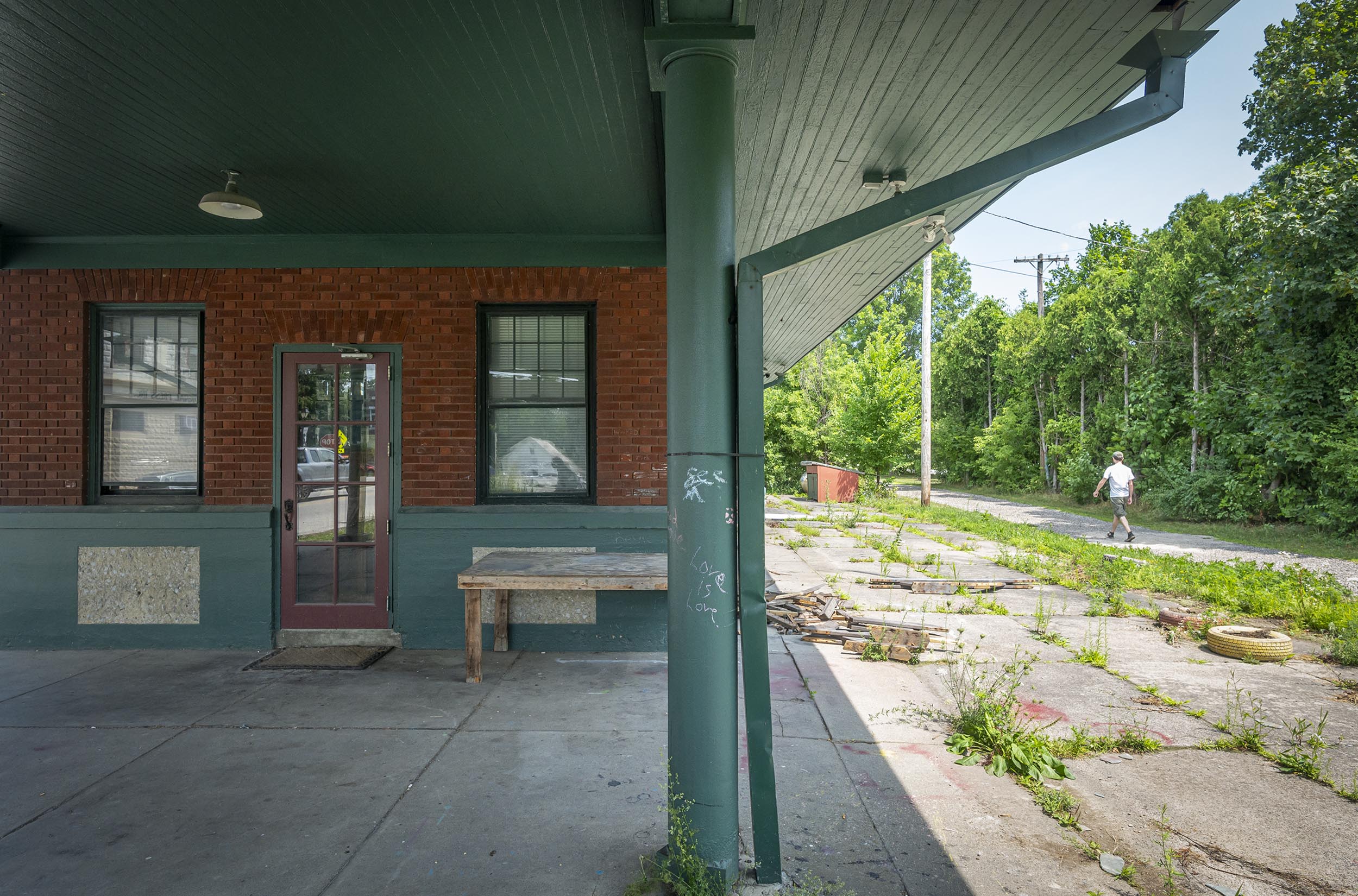 An abandoned building with boarded windows and a wooden table sits near a deserted pathway overgrown with weeds; a person is seen walking in the distance.