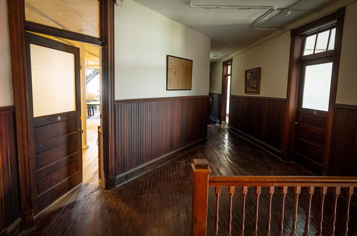 A dimly lit hallway with dark wooden floors and wainscoting, several partially open doors, and framed pictures on the walls.