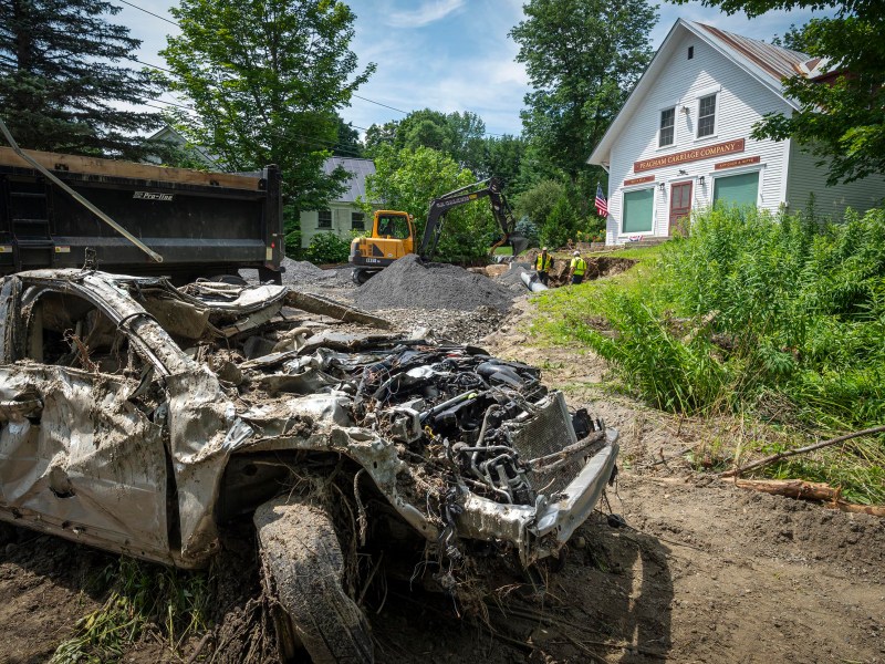 A damaged car sits in a yard while construction equipment works in the background near a white house surrounded by trees.
