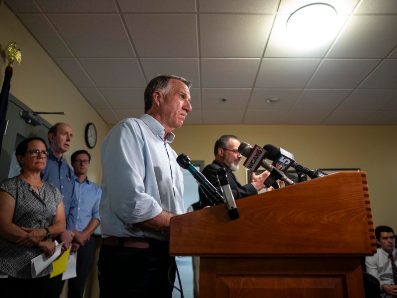 A man stands at a podium with microphones during a press conference, surrounded by individuals. The room has beige walls, a ceiling light, and a clock.