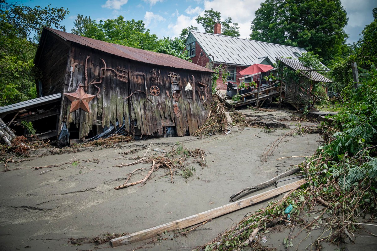 A dilapidated wooden structure, surrounded by debris and overgrown vegetation, appears to have been damaged, possibly by flooding or a landslide. In the background, a house with a metal roof is visible.