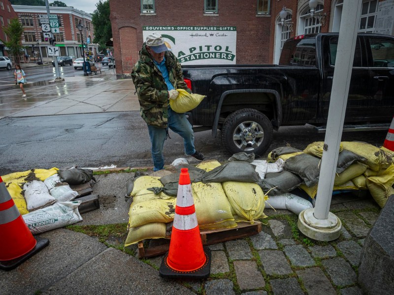 A man wearing a camouflage jacket and hat places sandbags on a wooden pallet near orange caution cones in front of a black truck and store on a rainy day in a city setting.