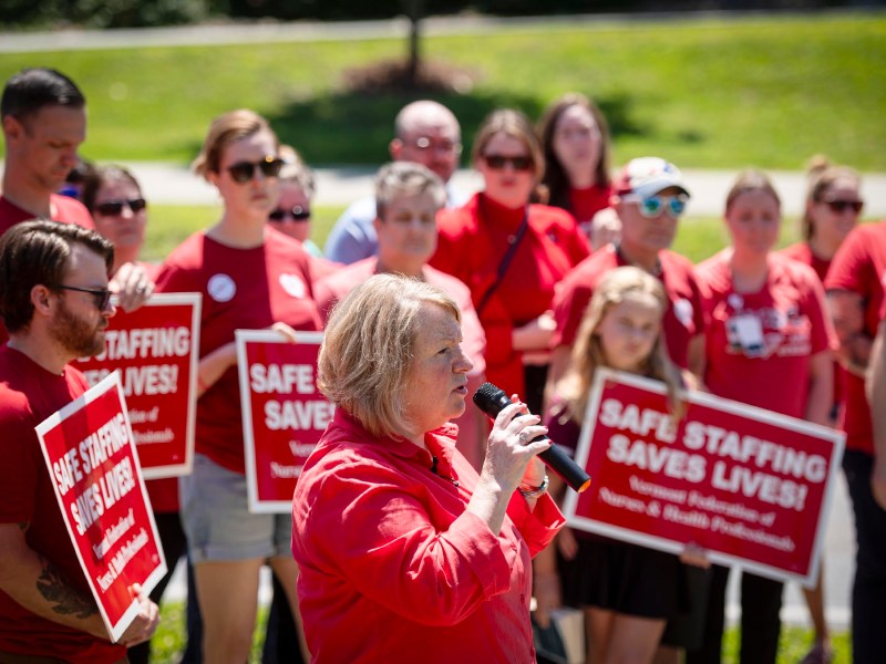 A group of people wearing red shirts are holding red and white signs that read "SAFE STAFFING SAVES LIVES!" A person in the foreground is speaking into a microphone.
