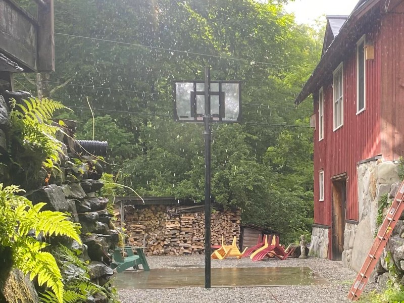 A basketball hoop stands on a gravel area between a red building and a stone wall. There is a pile of firewood in the background and a playground set to the right. It's raining heavily.