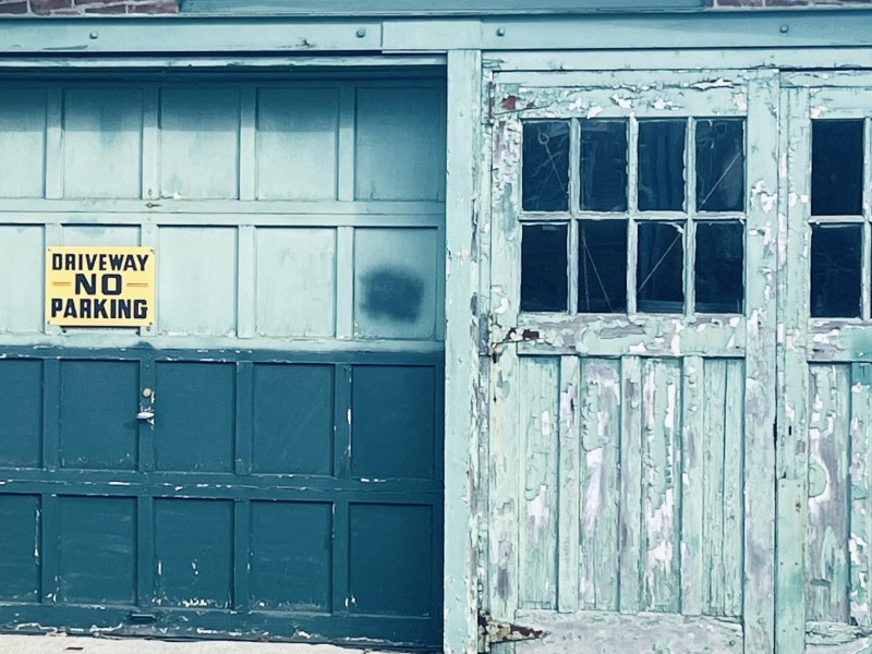 A weathered garage door with peeling teal paint and a "Driveway No Parking" sign. One side has glass window panels, some of which are broken.