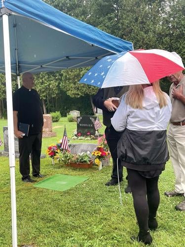 People gathered under a tent with umbrellas near a decorated gravesite; a clergyman is present. Two American flags and flowers are placed on the grave.